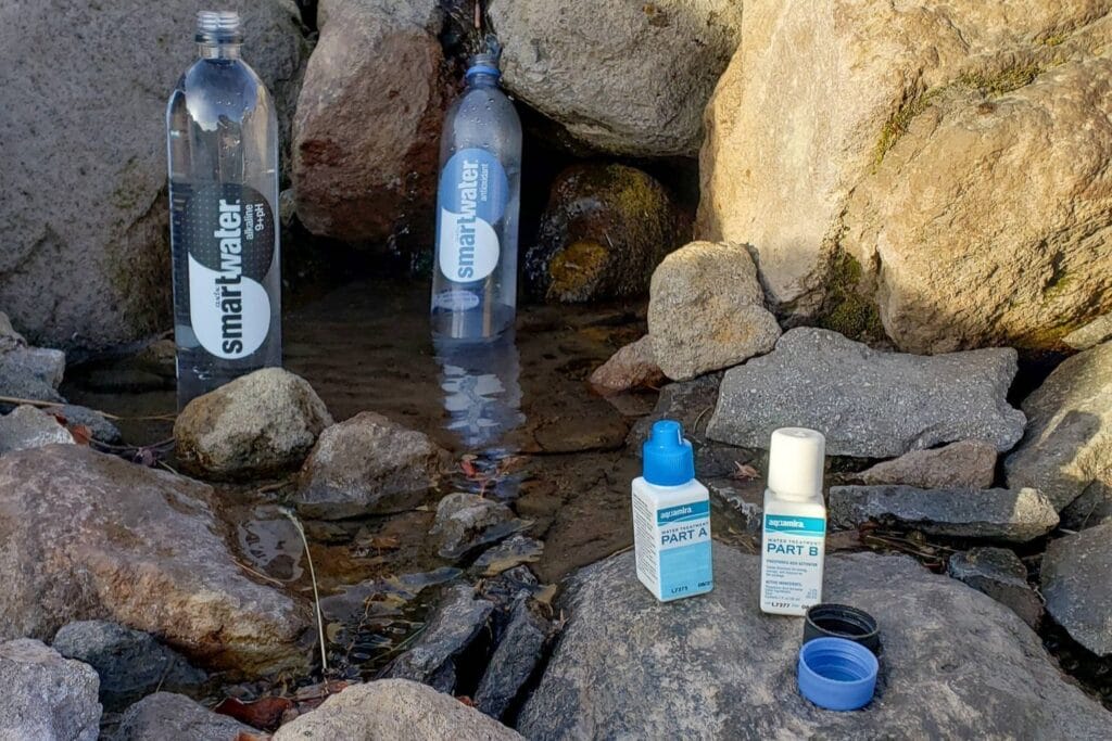 two water bottles in water with two bottles of Aquamira Water Treatment Drops lying on a stone