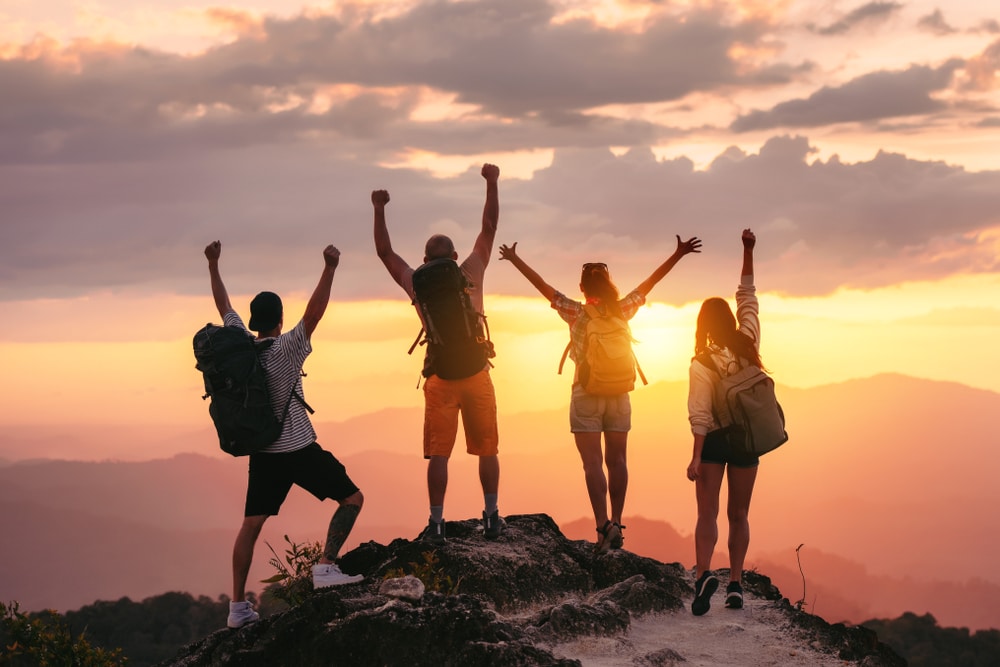 Happy friends hikers or tourists stands with raised arms on mountain top against mountains and looking at sunset