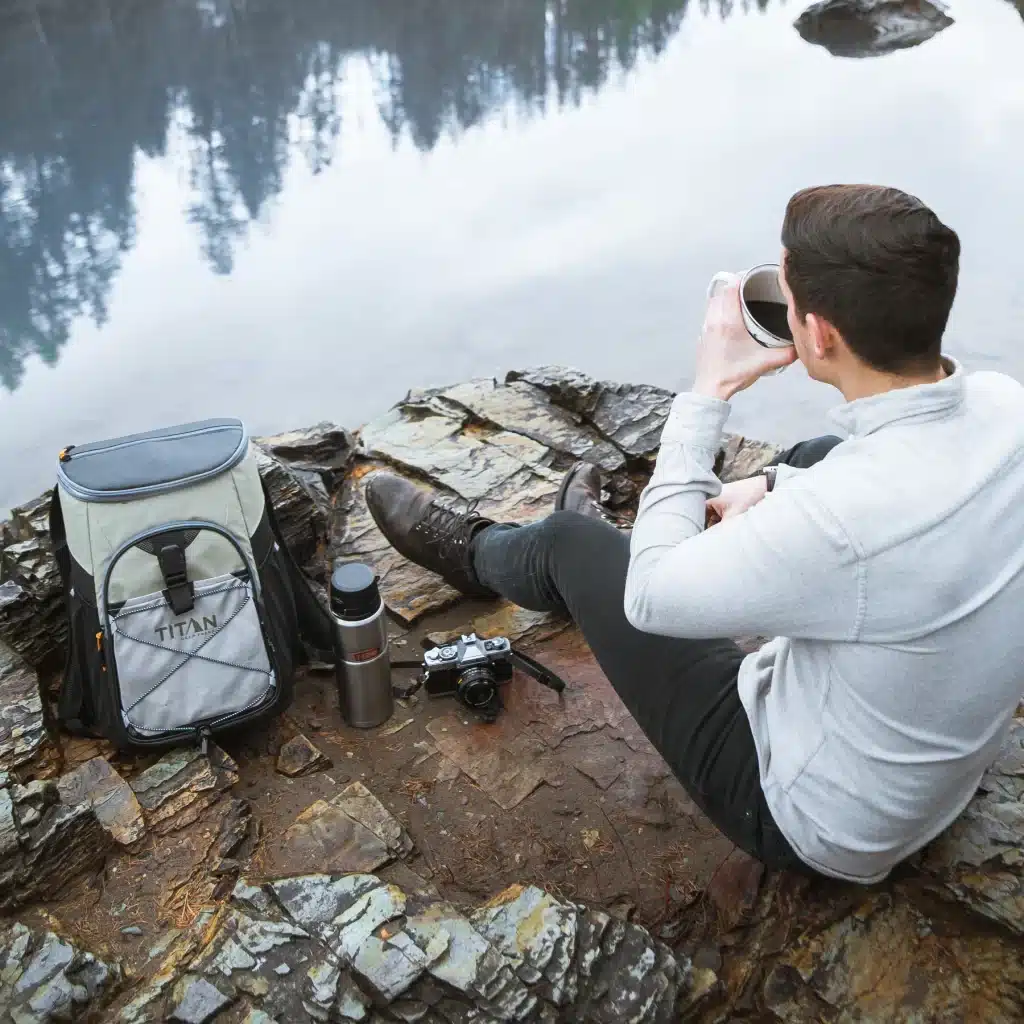 a man with his Arctic Zone Titan Deep Freeze Backpack Cooler sitting along riverside