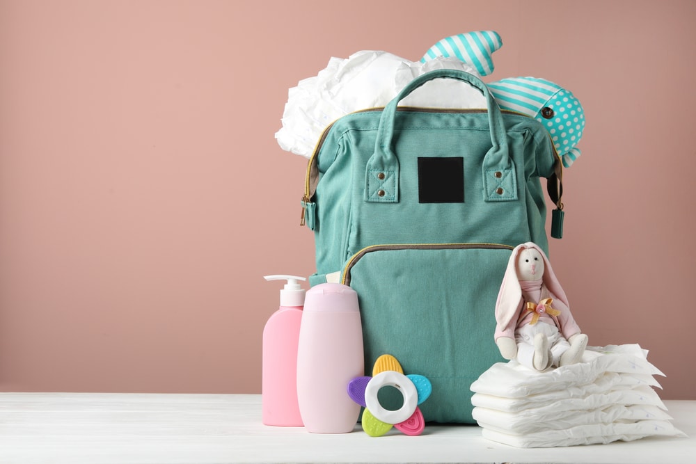 Bag with diapers and baby accessories on white wooden table against pink background