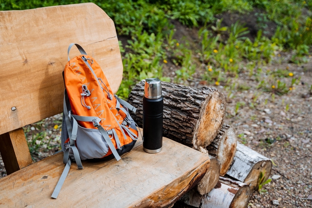 Orange And Gray Daypack With Thermos Resting On A Wooden bench